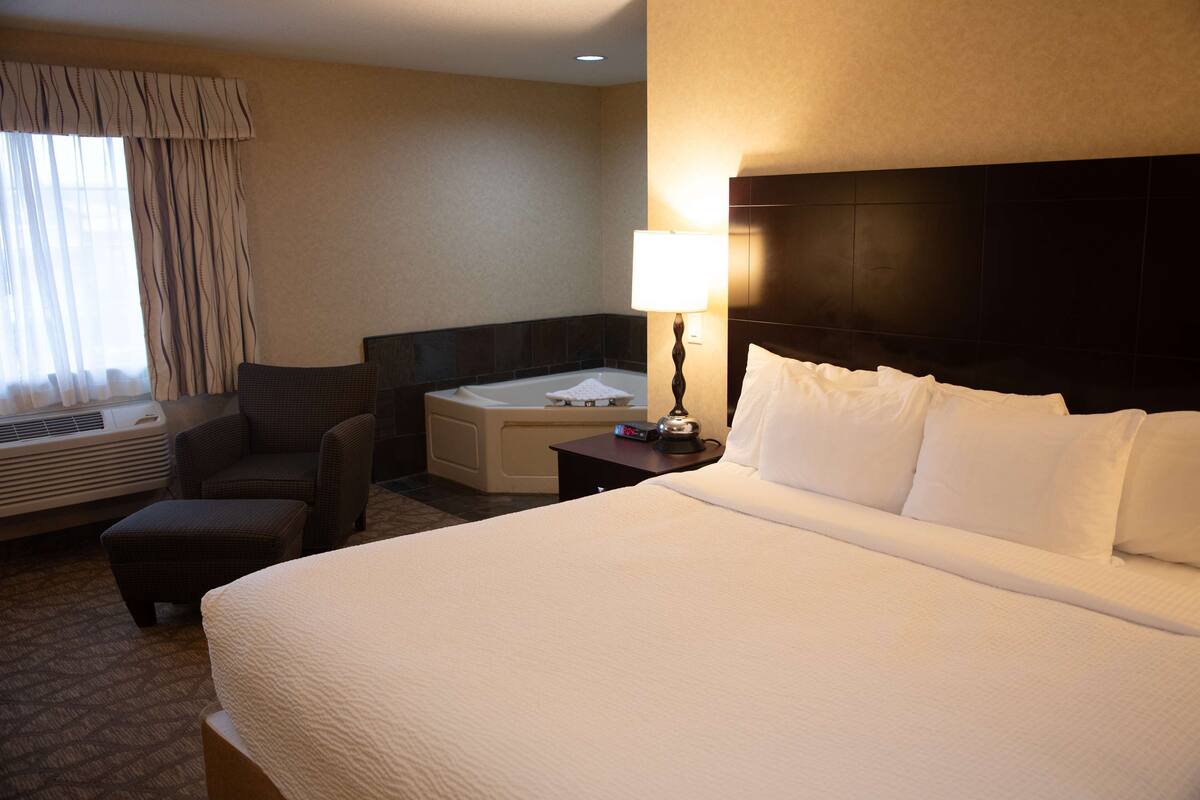 A hotel room with a large bed, a sitting area with a chair and ottoman, a bathtub, and a wall-mounted lamp.