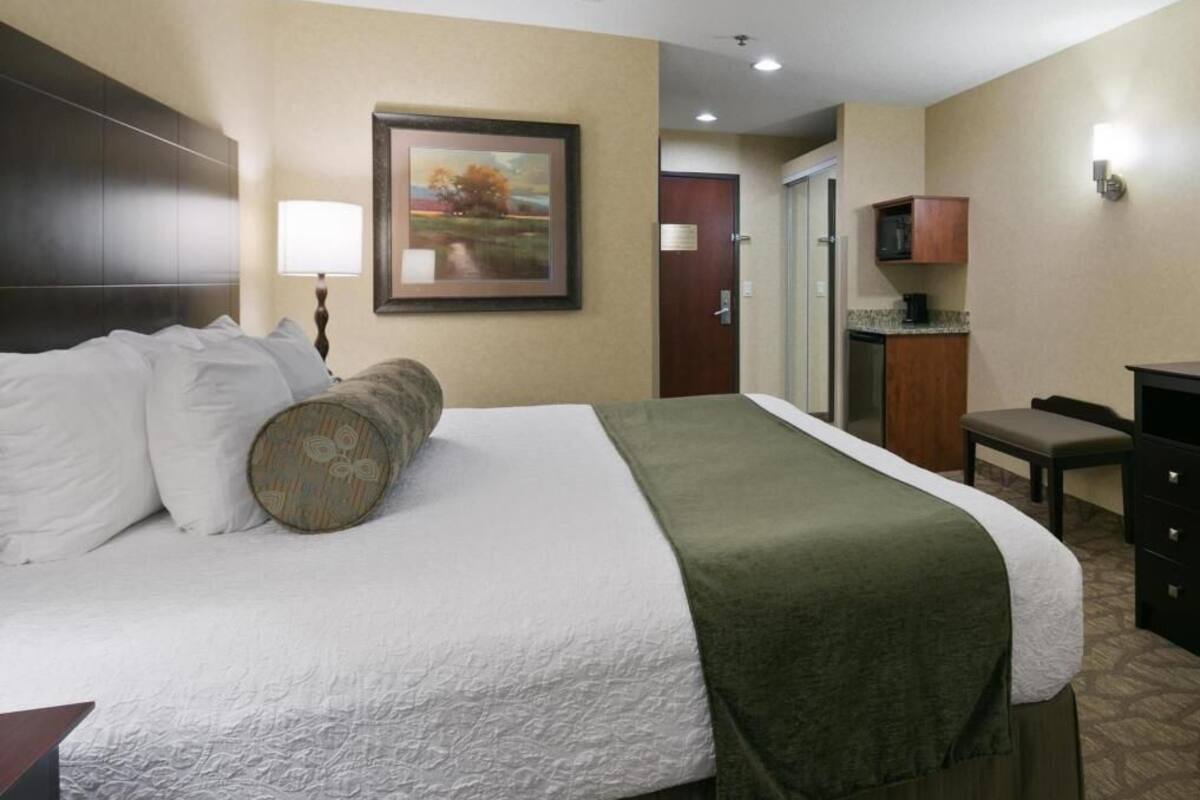 A hotel room with a large bed, a desk, a chair, a lamp, a painting on the wall, and a door leading to another room.