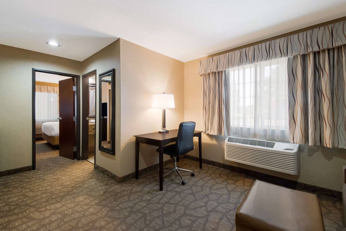 A hotel room with a desk, chair, and an air conditioning unit. There is a mirror on the wall and a door leading to another room.