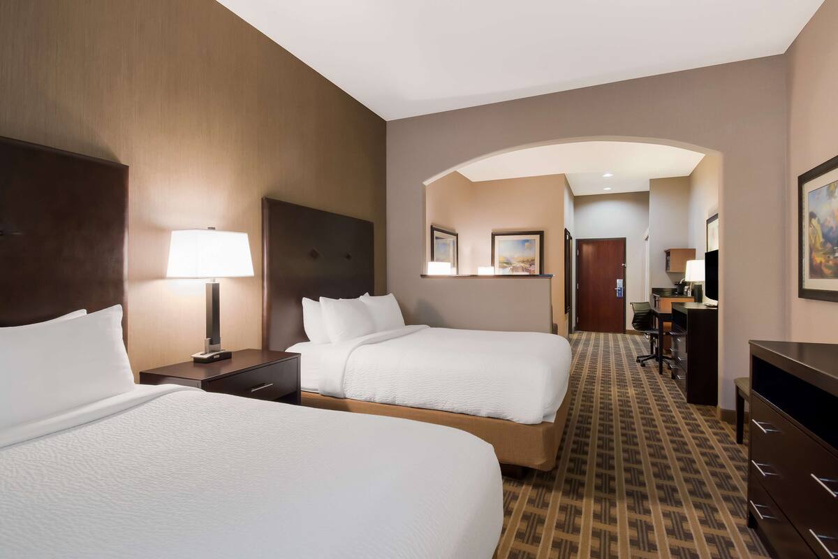 A hotel room with two beds, a desk, a lamp, and a view of the hallway.