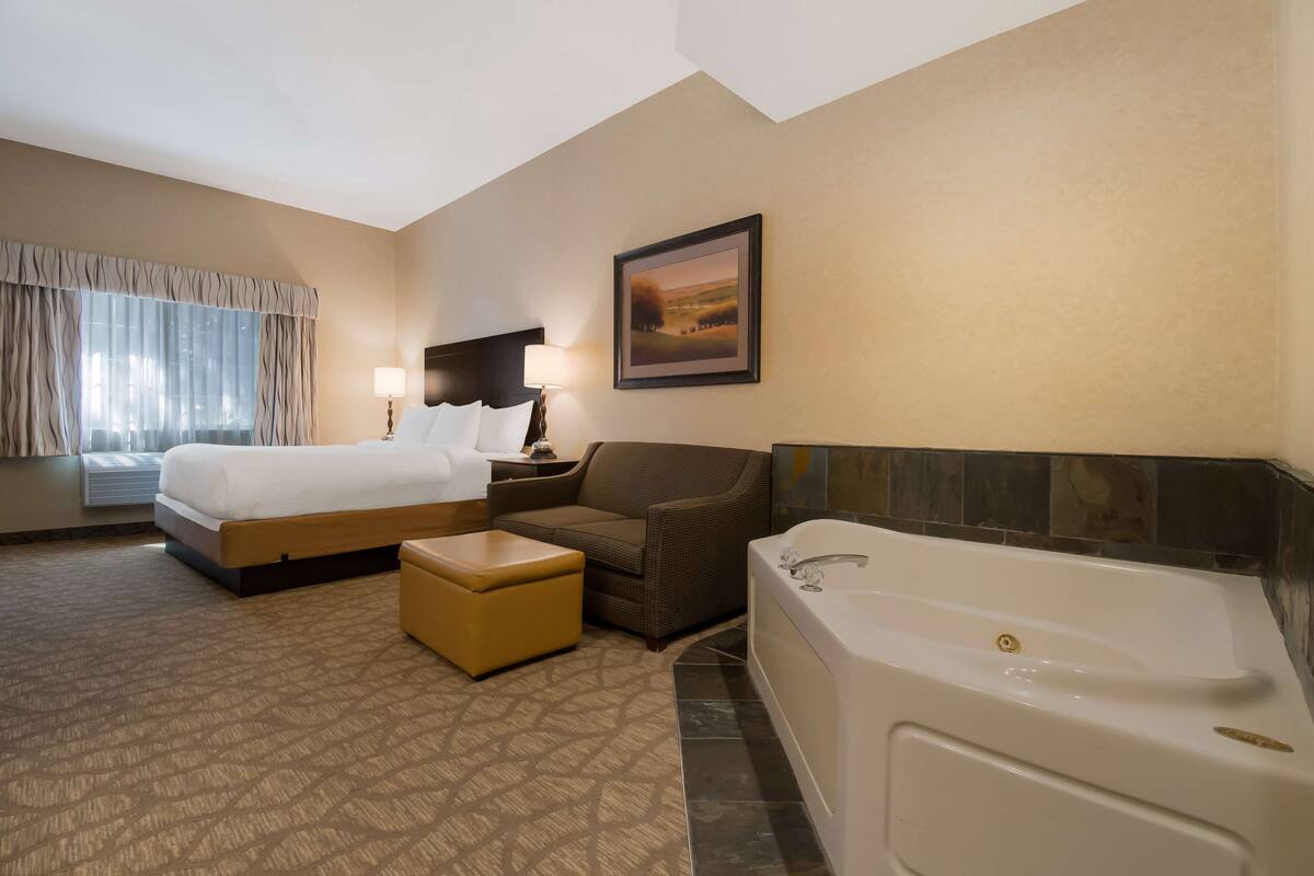 A hotel room with a bed, a sofa, a small ottoman, a bathtub, and a painting on the wall.
