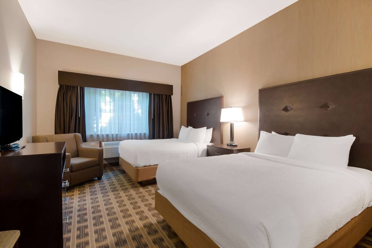 A hotel room with two beds, a desk, a chair, a TV, and a large window with curtains.