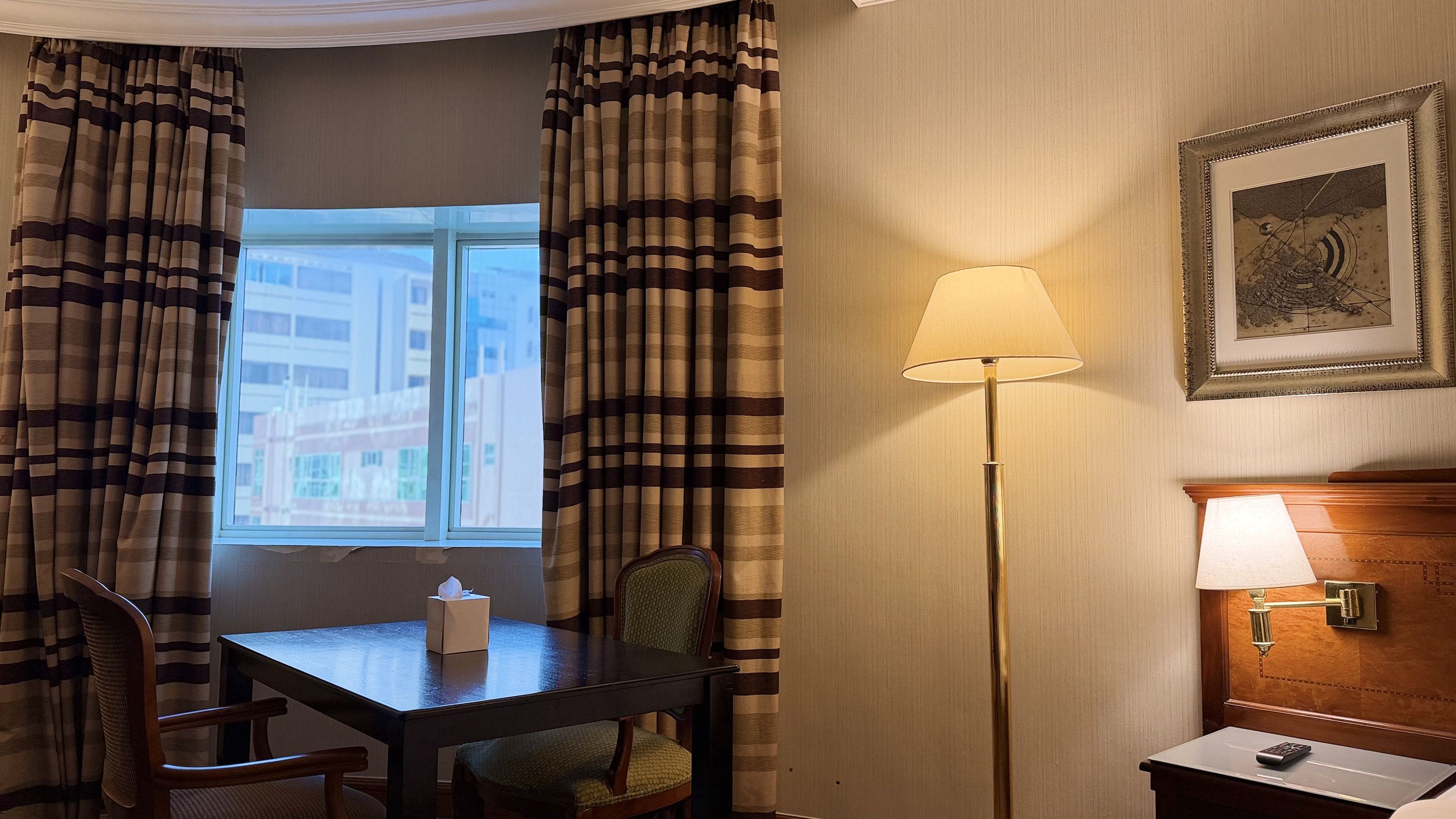 Ein Hotelzimmer mit Tisch, Stühlen, einer Lampe, einem gerahmten Bild an der Wand und Vorhängen.