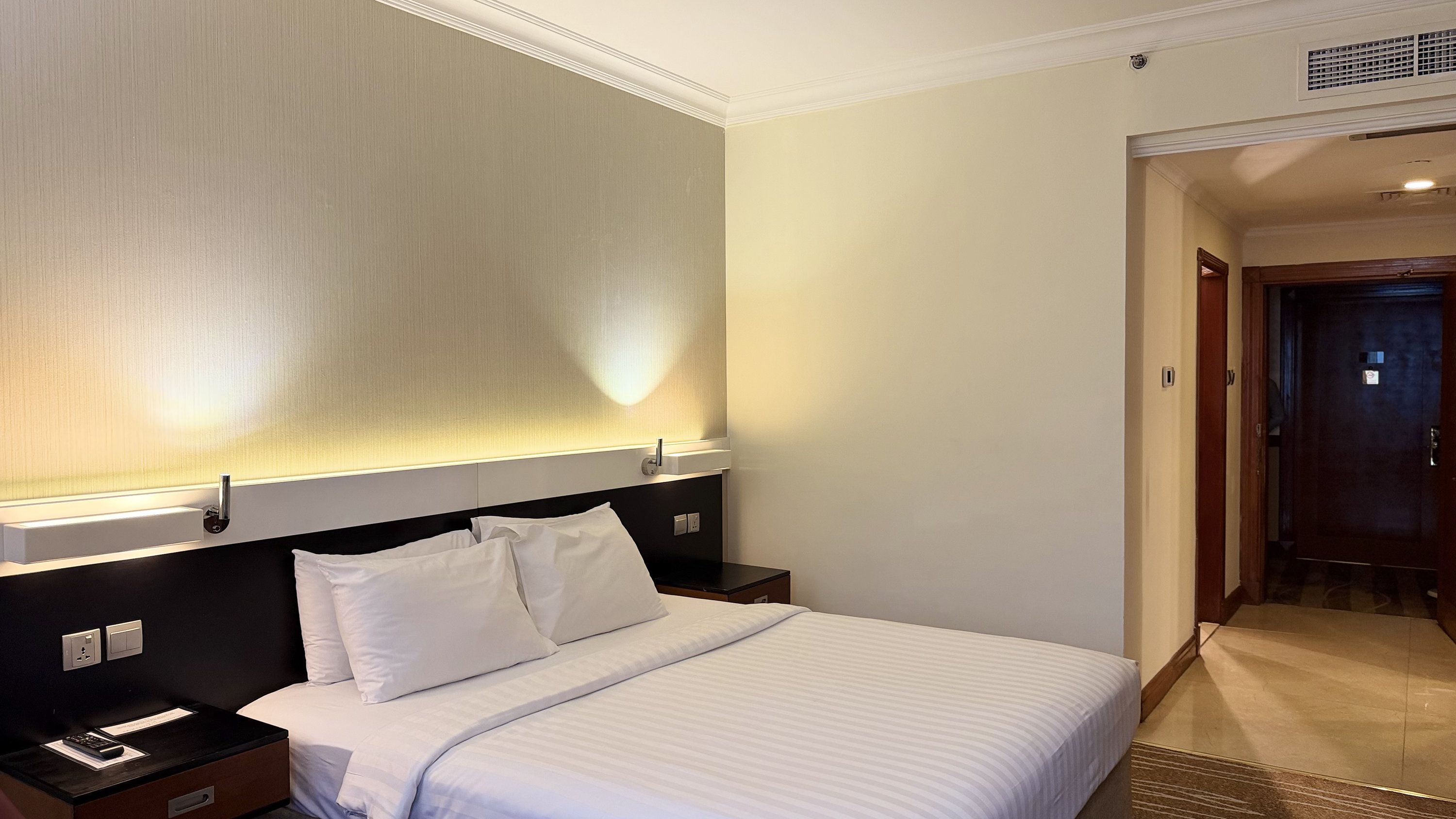 A hotel room with a bed, bedside tables, a nightstand, a wall-mounted light, and a door.