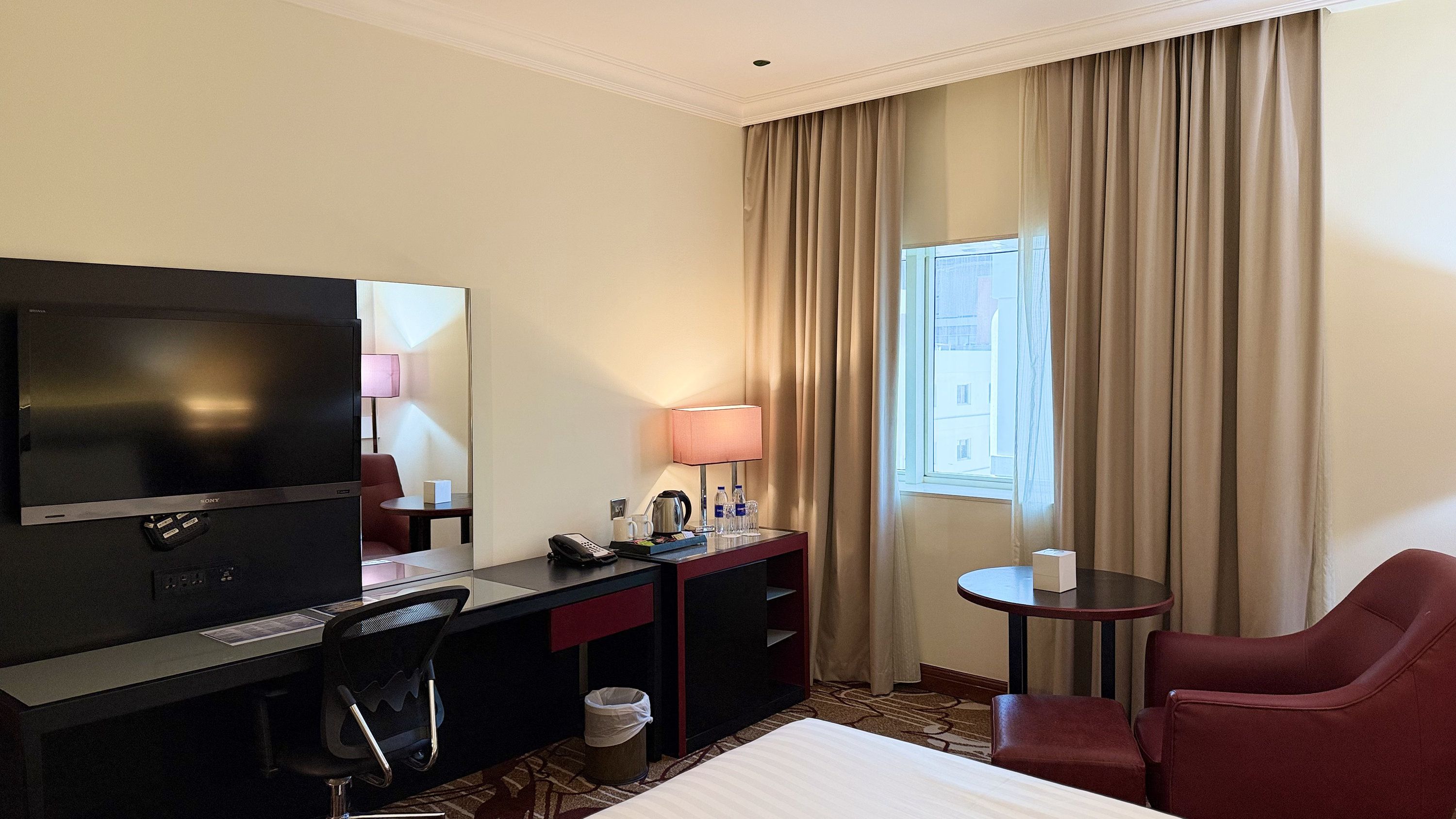 A hotel room with a bed, a desk with a television, a lamp, a chair, and a window with curtains.