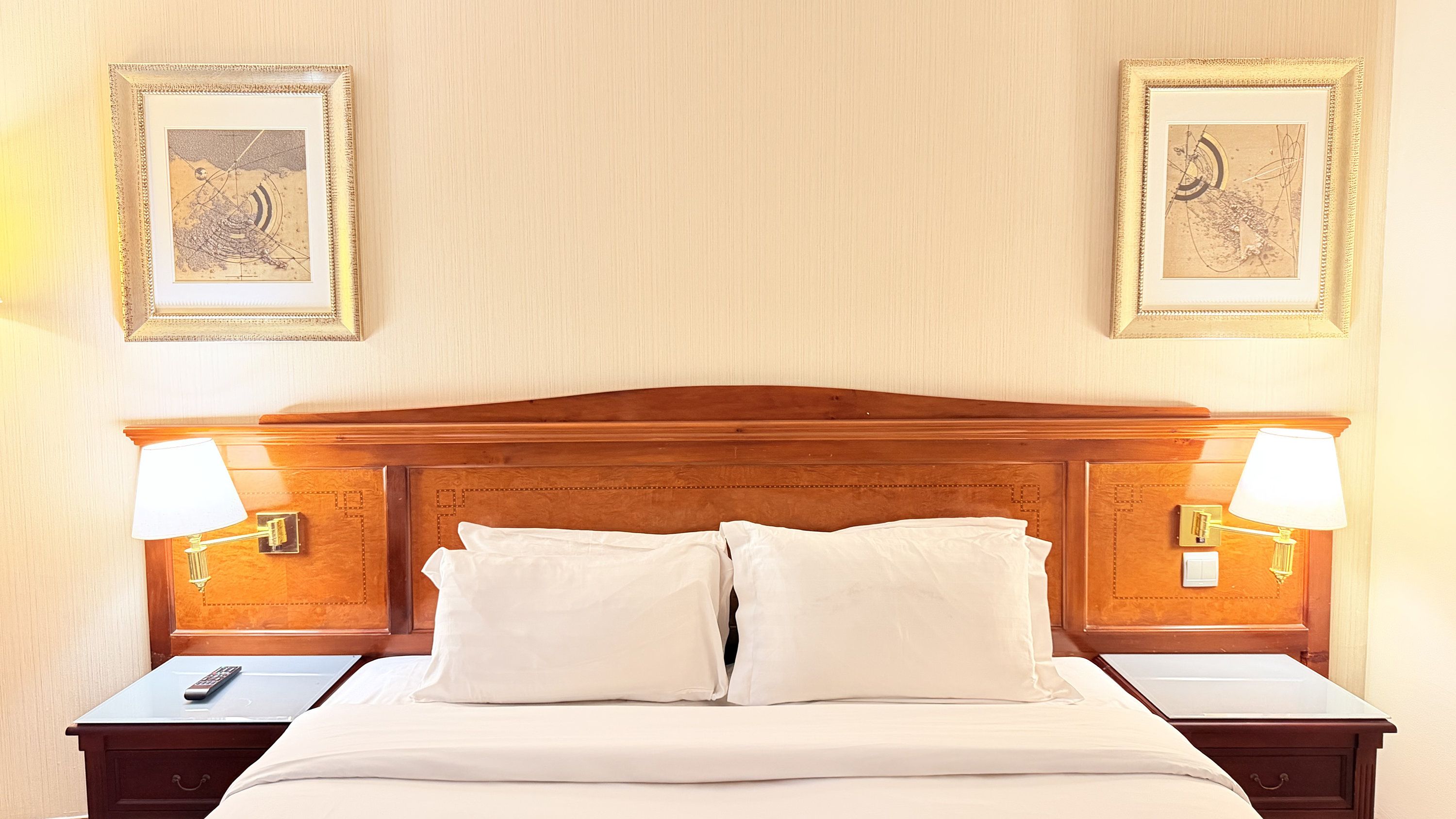 Een hotelkamer met een houten hoofdeinde, twee bedlampjes, een bed met witte lakens en twee ingelijste foto's aan de muur.