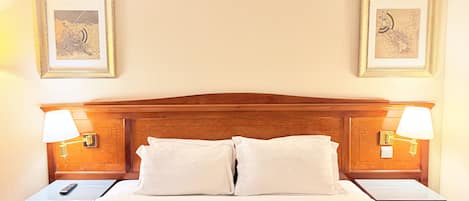 A hotel room with a wooden headboard, two bedside lamps, a bed with white linens, and two framed pictures on the wall.