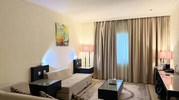 Habitación de hotel con sofá, mesa de centro, escritorio con lámpara, un cuadro en la pared y un ventanal con cortinas.