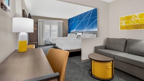A hotel room with a bed, a sofa, a dining table, a lamp, and a wall art piece. - Comfort Suites Fort Wayne North (Fort Wayne)