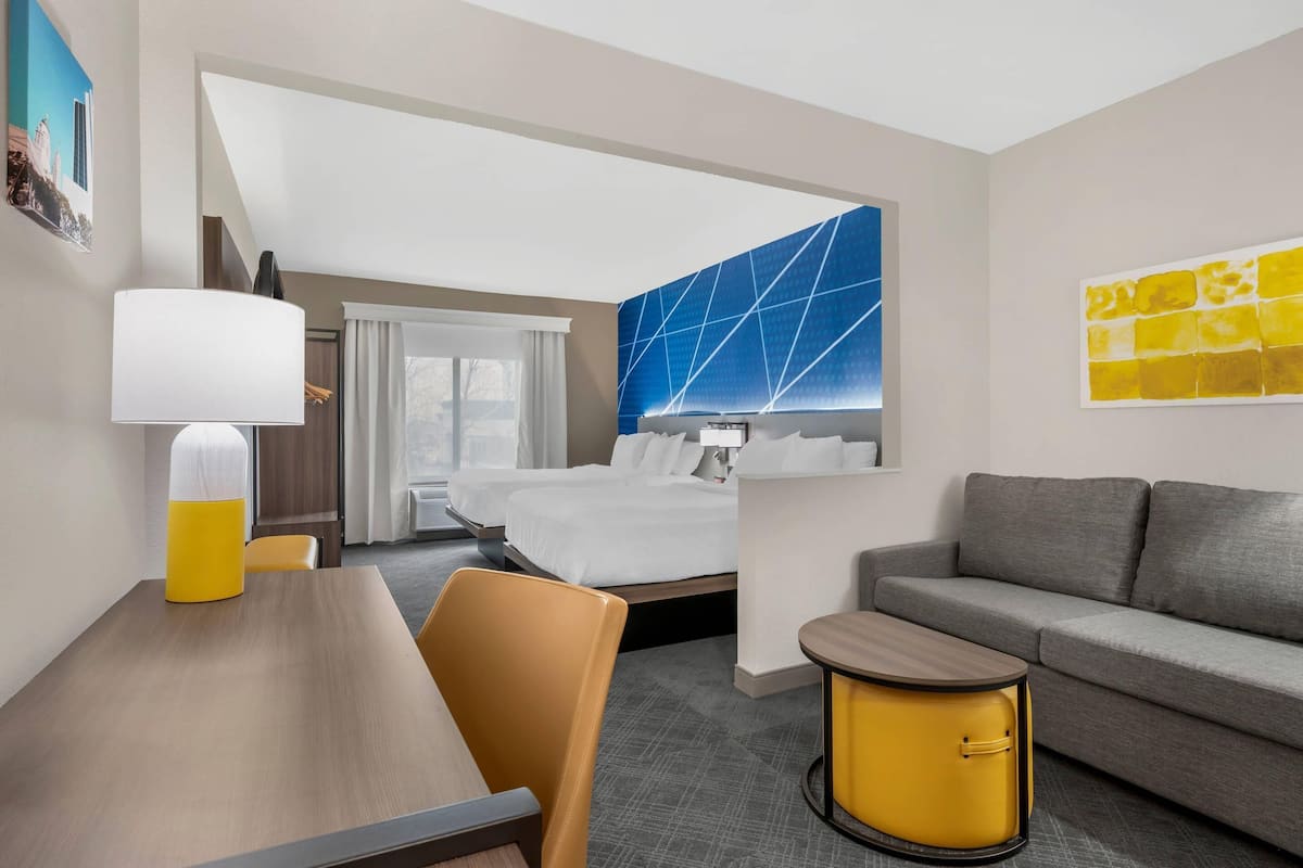 A hotel room with a bed, a sofa, a dining table, a lamp, and a wall art piece.