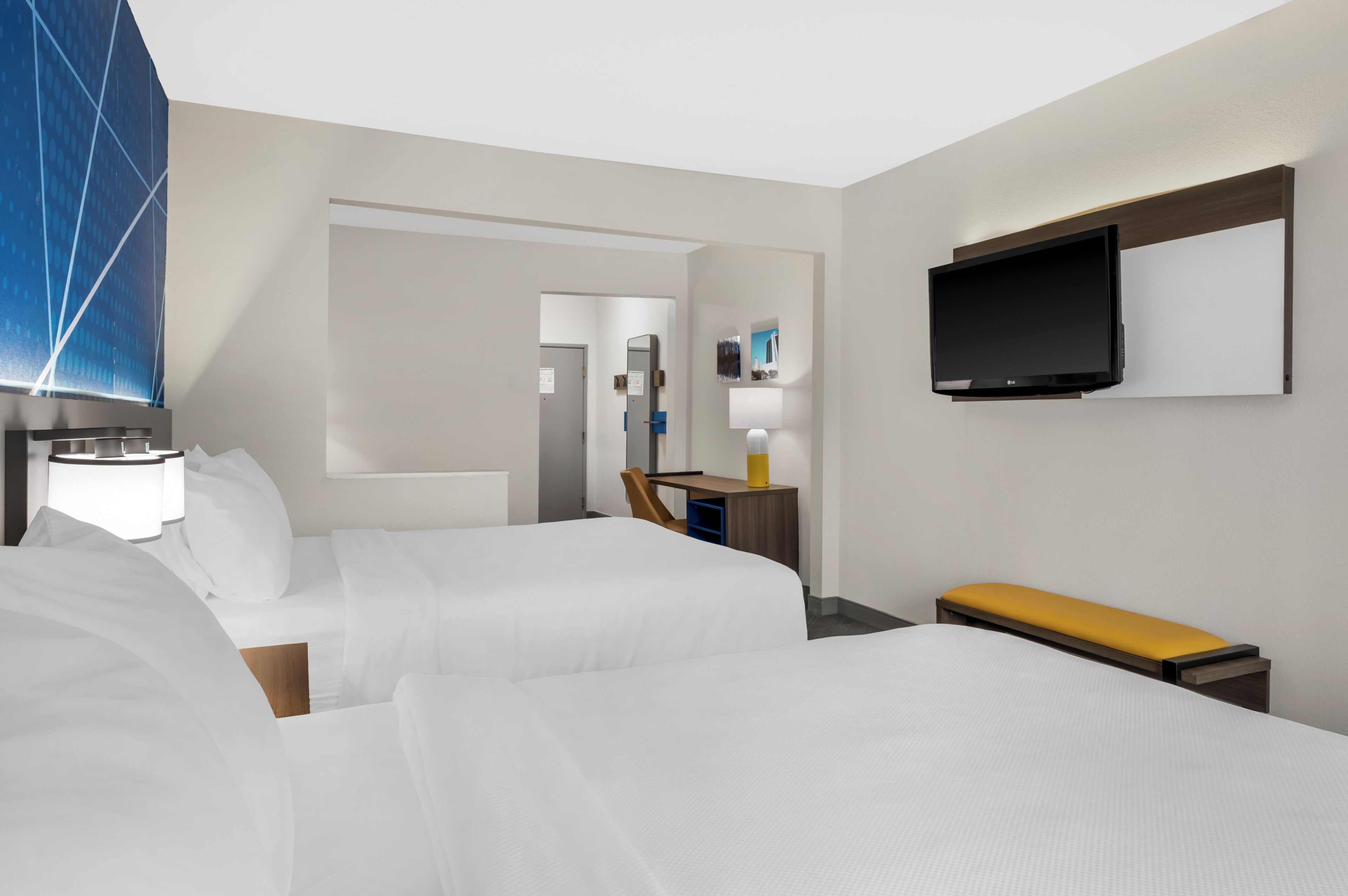 A hotel room with two beds, a TV, a desk with a chair, and a bathroom visible through an open door.
