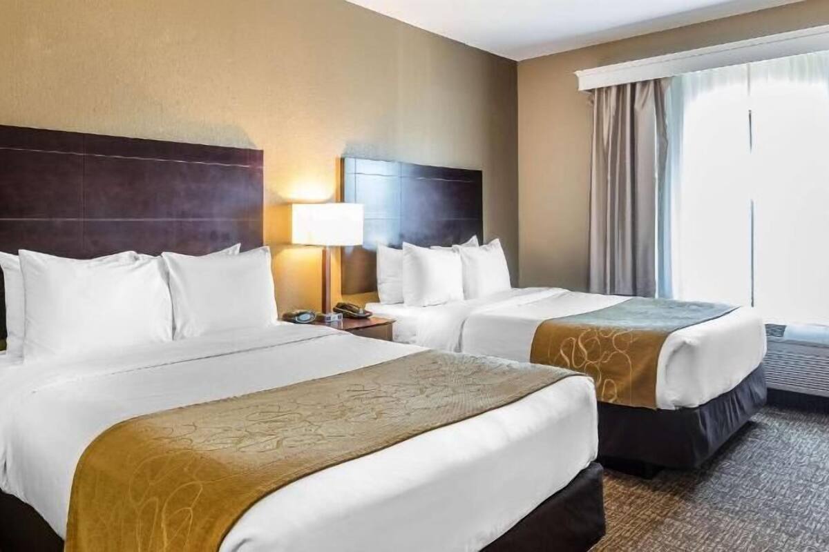 A hotel room with two beds, a nightstand, a lamp, and a large window with curtains.