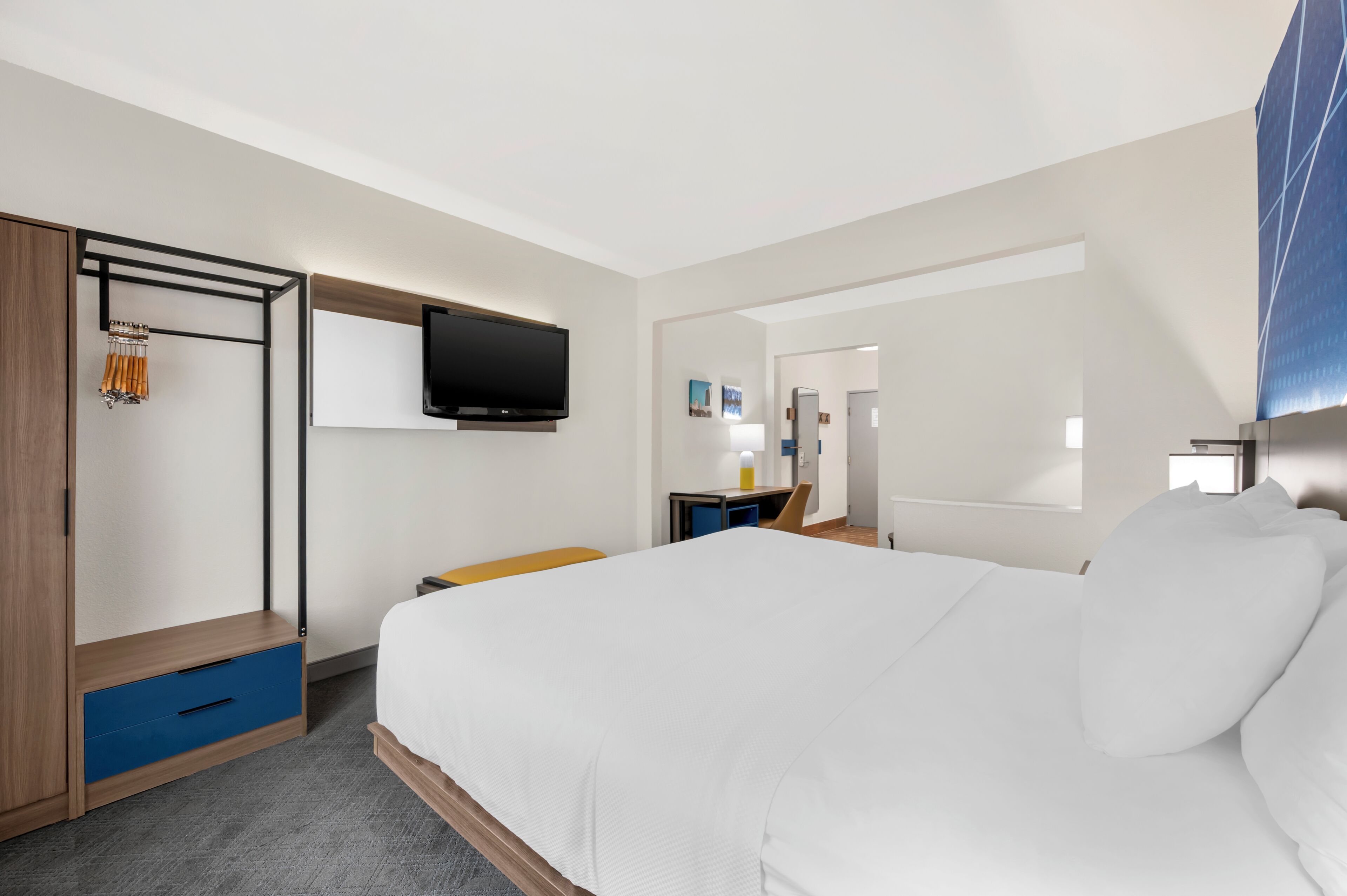 A hotel room with a bed, a TV mounted on the wall, a desk with a chair, and a wardrobe with a mirror.