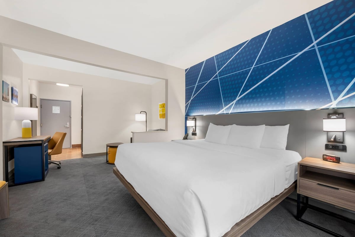A hotel room with a large bed, a desk, a chair, a nightstand, and a wall-mounted TV.