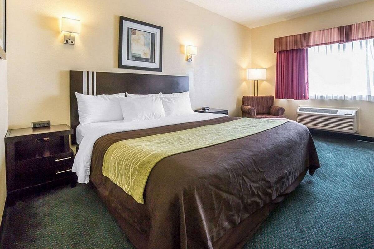 A hotel room with a large bed, two bedside tables, a chair, a lamp, an air conditioning unit, and a window with curtains.