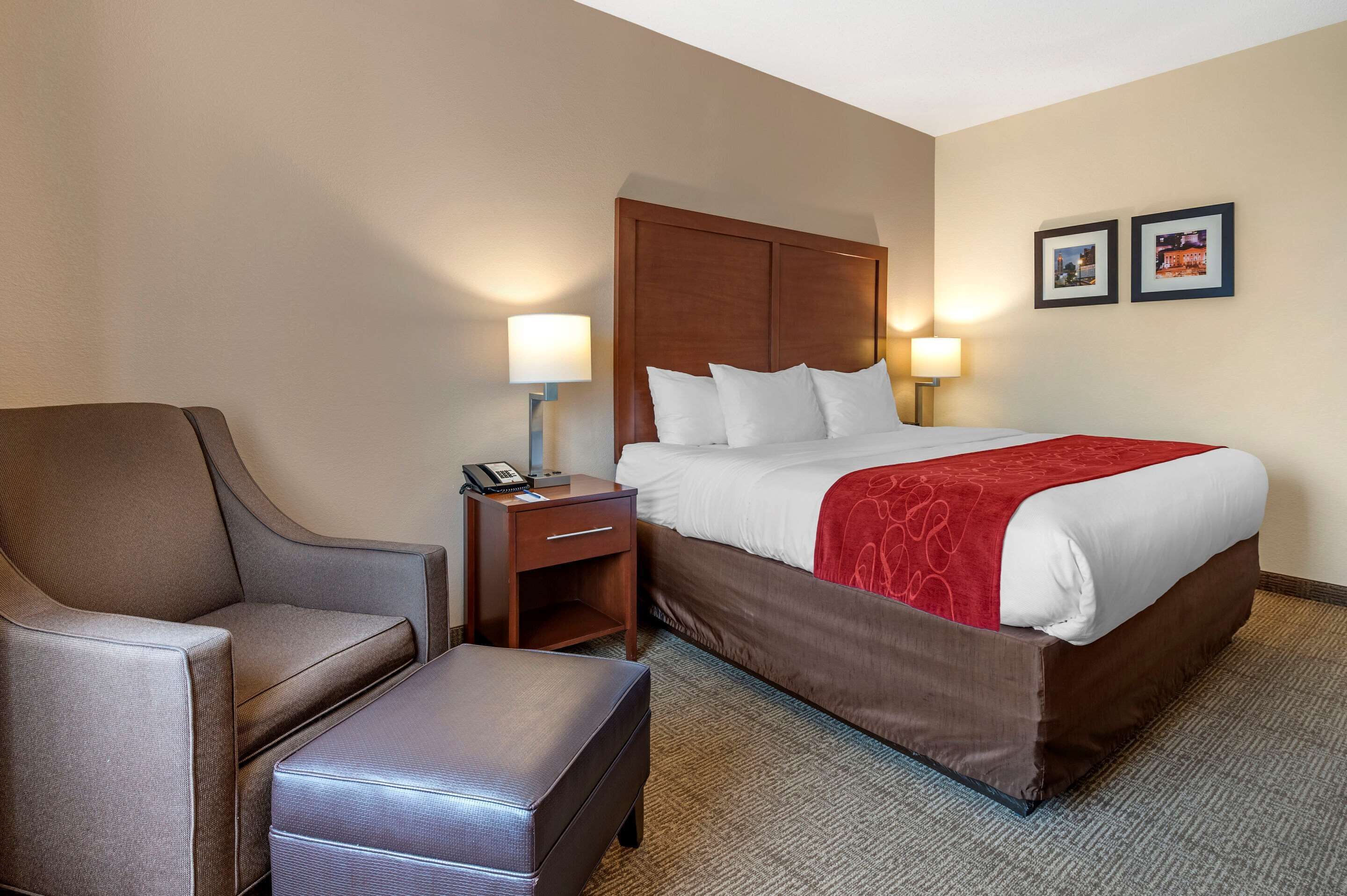 A hotel room with a bed, a chair, a nightstand with a lamp, and a small table.