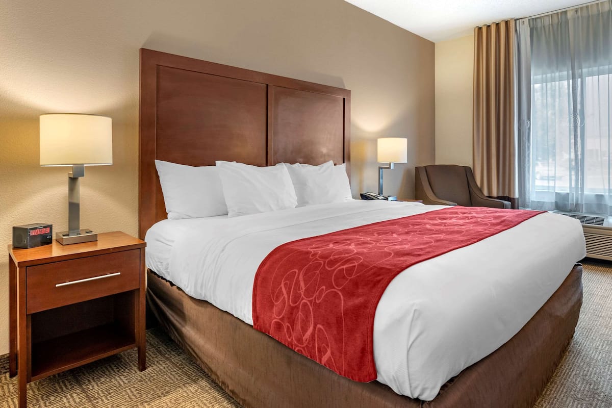 A hotel room with a large bed, a wooden headboard, two bedside tables with lamps, a chair, and a window with curtains.