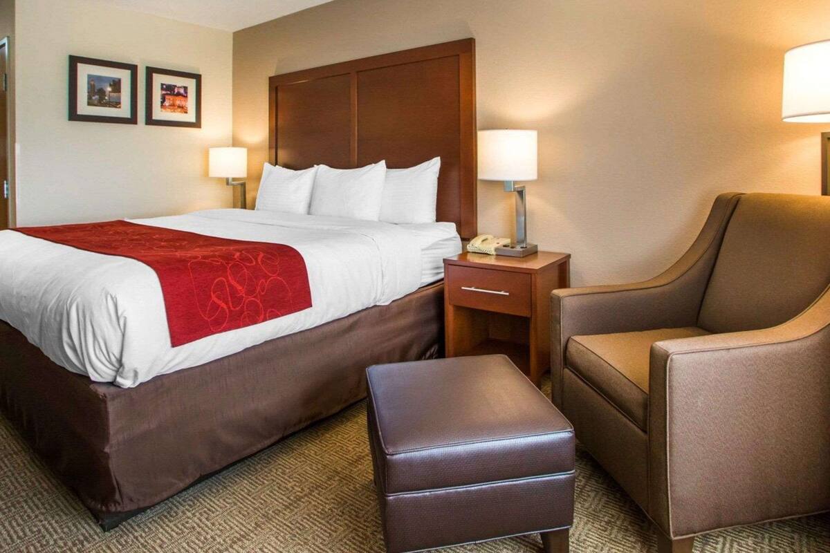 A hotel room with a large bed, two bedside lamps, a wooden headboard, a brown leather armchair, and a small ottoman.