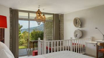 A bedroom with a large bed, a desk, a chair, a balcony with a table and chairs, and a view of greenery and mountains.