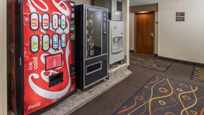 Een hal met een Coca-Cola automaat voor ijskoud drank, een drankautomaat en een automaat met snacks. Achtergrond toont een deur.