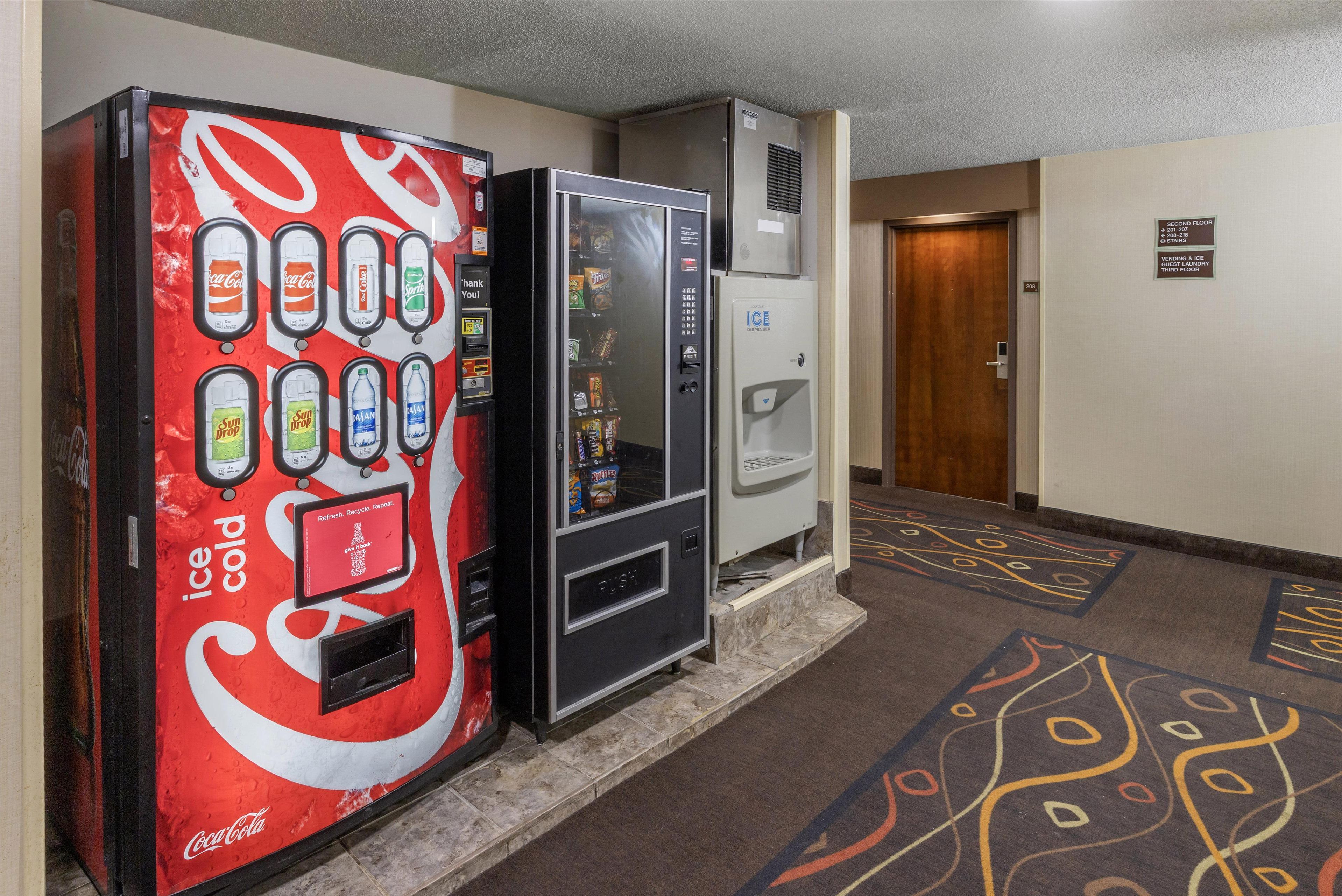 Un pasillo con una máquina expendedora Coca-Cola que ofrece bebidas heladas, un dispensador de bebidas y una máquina expendedora de snacks. Se ve una puerta al fondo.