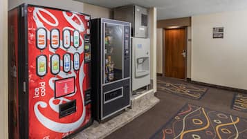 Un pasillo con una máquina expendedora Coca-Cola que ofrece bebidas heladas, un dispensador de bebidas y una máquina expendedora de snacks. Se ve una puerta al fondo.