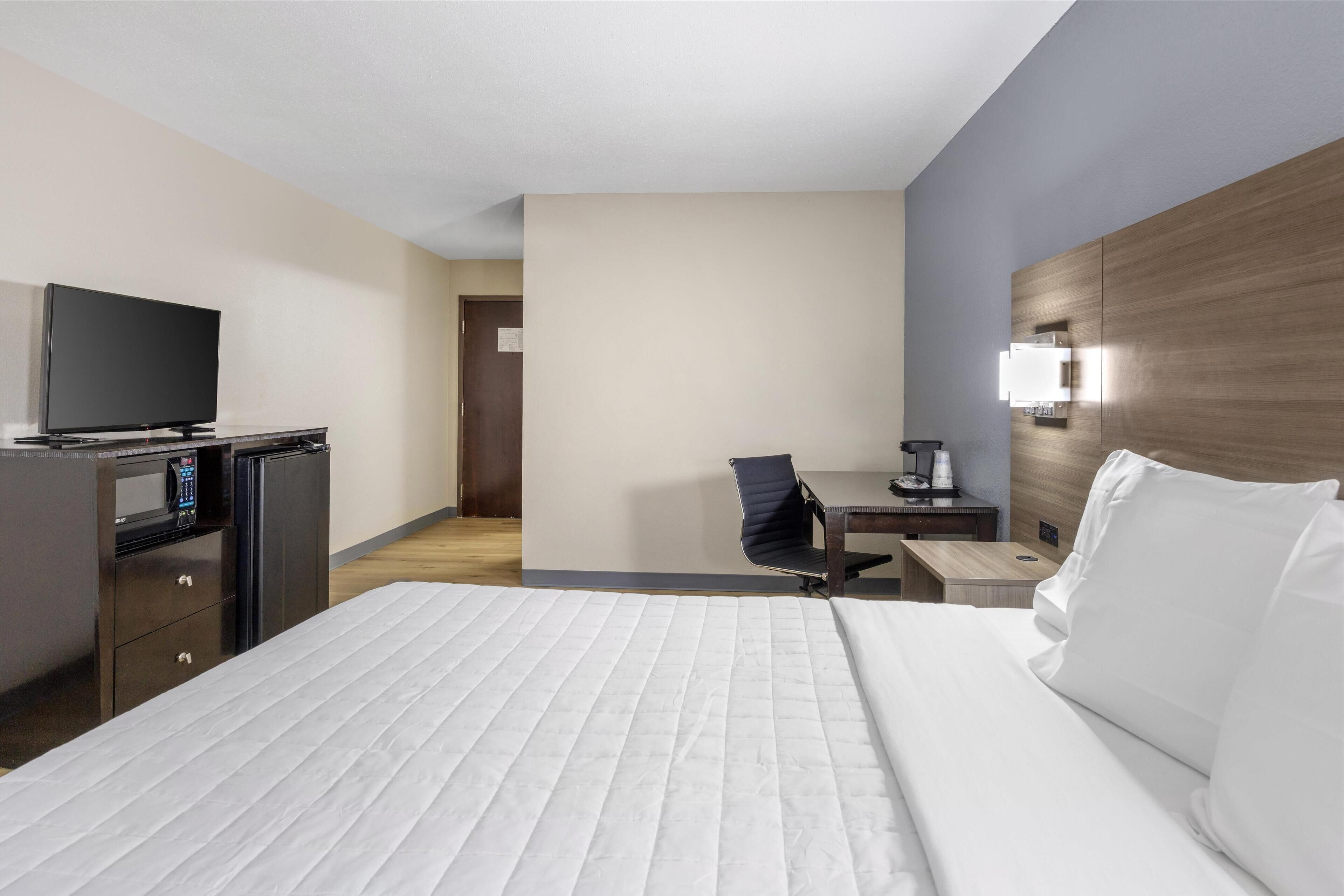 A hotel room with a bed, a desk with a chair, a television, a microwave, and a nightstand with a lamp.