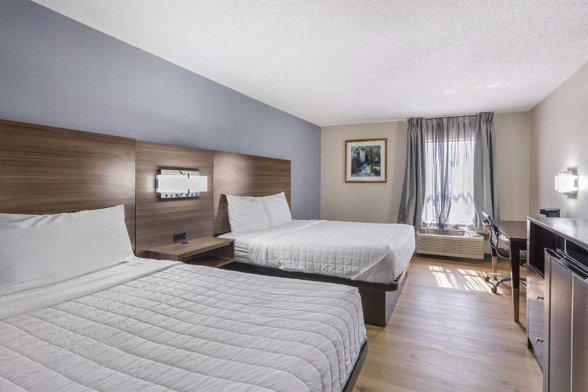 Ein Hotelzimmer mit zwei Betten, einem Schreibtisch, einem Stuhl und einem fensterbepflanzten Fenster.