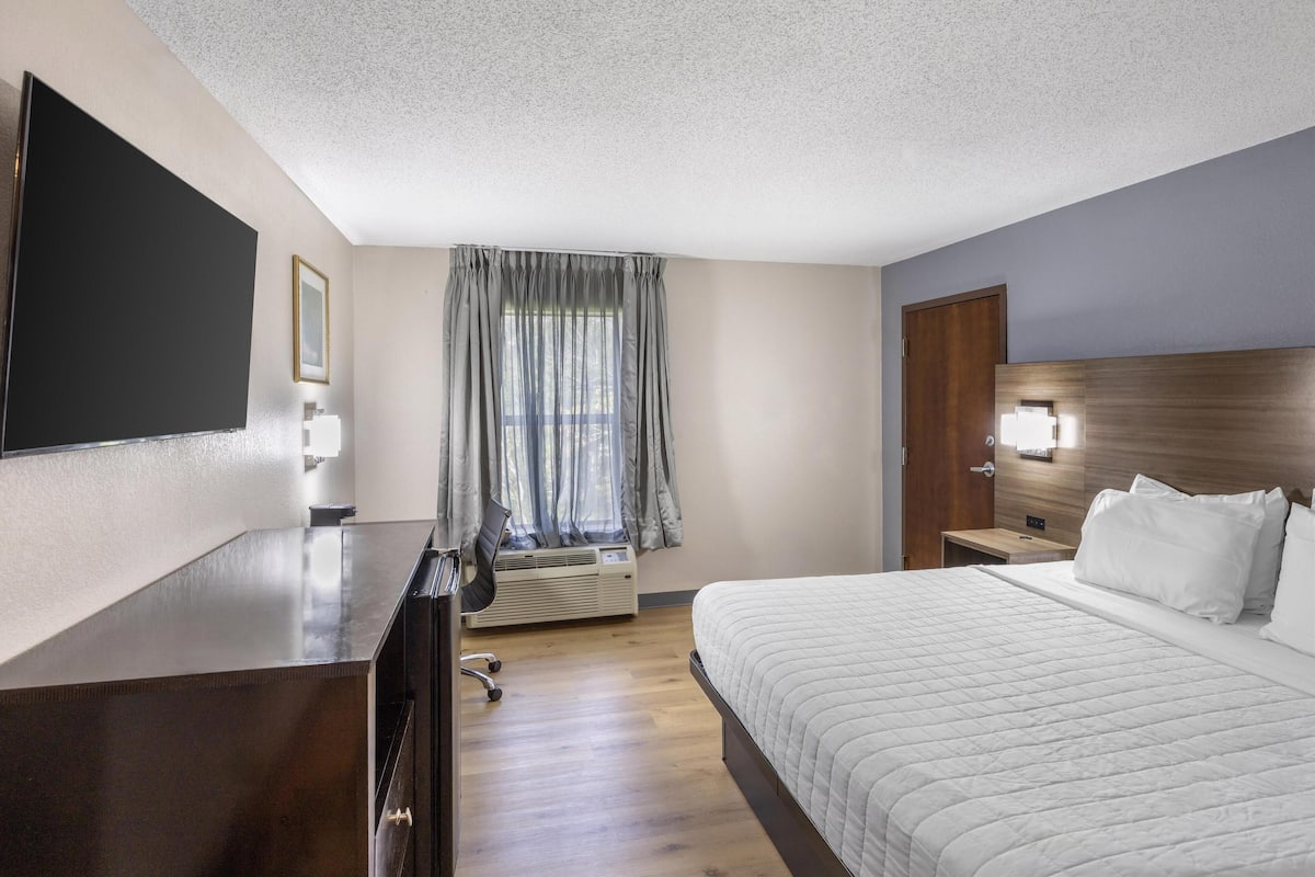 Ein Hotelzimmer mit einem großen Bett, einem Schreibtisch mit Stuhl, einem Fernseher, einem Fenster mit Vorhängen und einem hölzernen Kopfteil.
