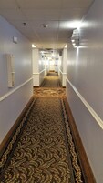 Un pasillo de hotel con alfombra estampada, paredes blancas y una puerta al final.
