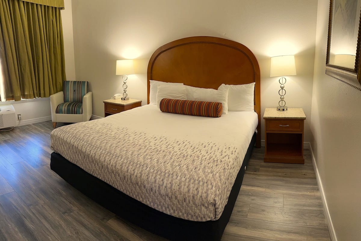 Habitación de hotel con una cama grande, dos mesitas de noche con lámparas, una silla y un ventanal con cortinas.