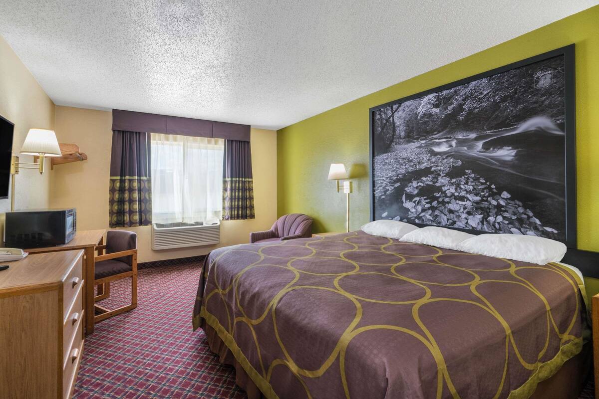 A hotel room with a large bed, a desk with a computer, a chair, a TV, and a large window with curtains.