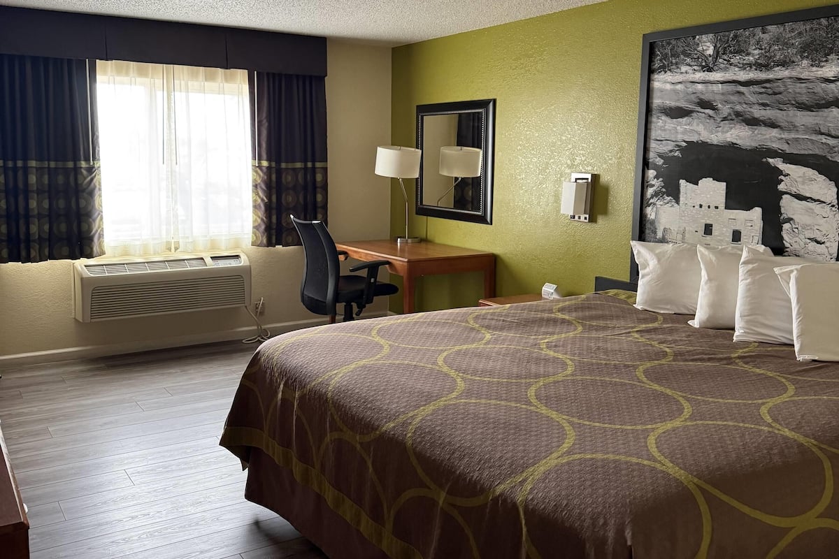 A hotel room with a large bed, a desk with a chair, a window with curtains, an air conditioning unit, a wall-mounted mirror, and a framed picture.