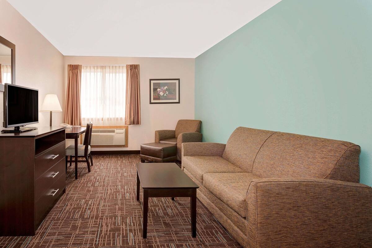 Hotelkamer met een bruine bank, een klein tafeltje, een televisie op een houten kast, een stoel, een lamp, een raam met gordijnen en een ingelijst schilderij aan de muur.