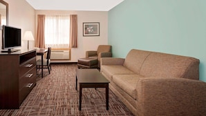 Habitación de hotel con un sofá marrón, una mesita, un televisor sobre un mueble de madera, una silla, una lámpara, una ventana con cortinas y un cuadro enmarcado en la pared.