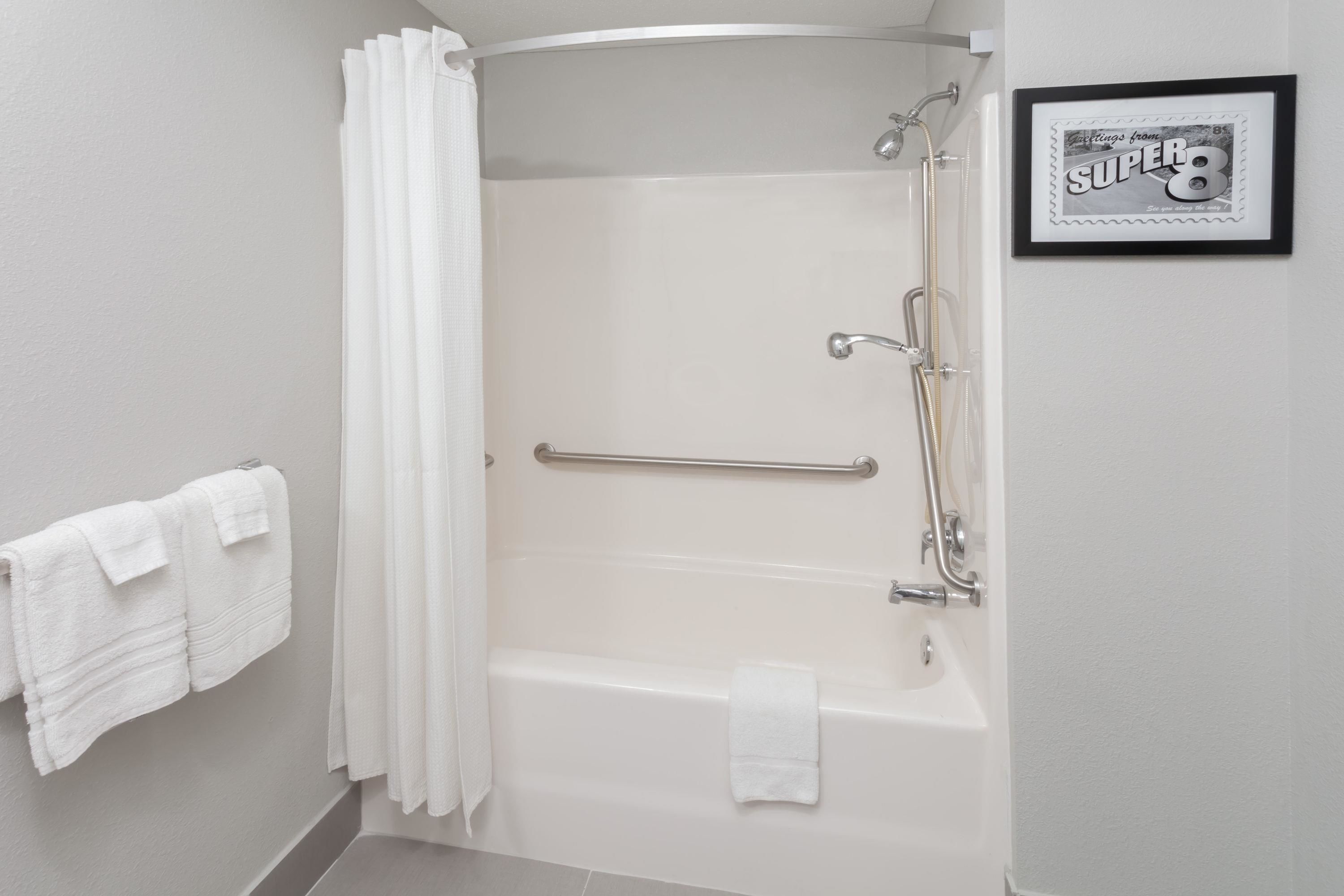 A modern bathroom with a bathtub, a shower, and a handrail. There is a framed picture on the wall and a towel rack with towels.
