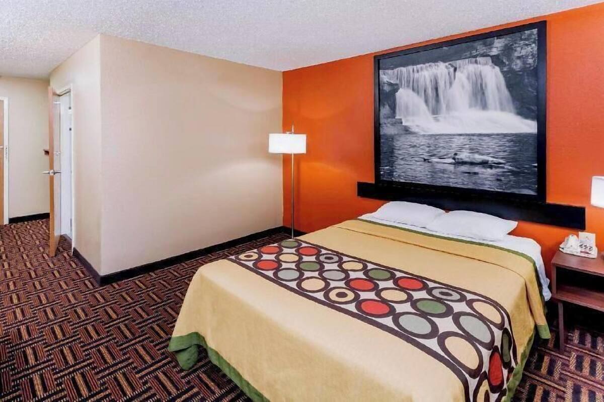 A hotel room with a bed, a wall-mounted TV, a lamp, and a patterned carpet.