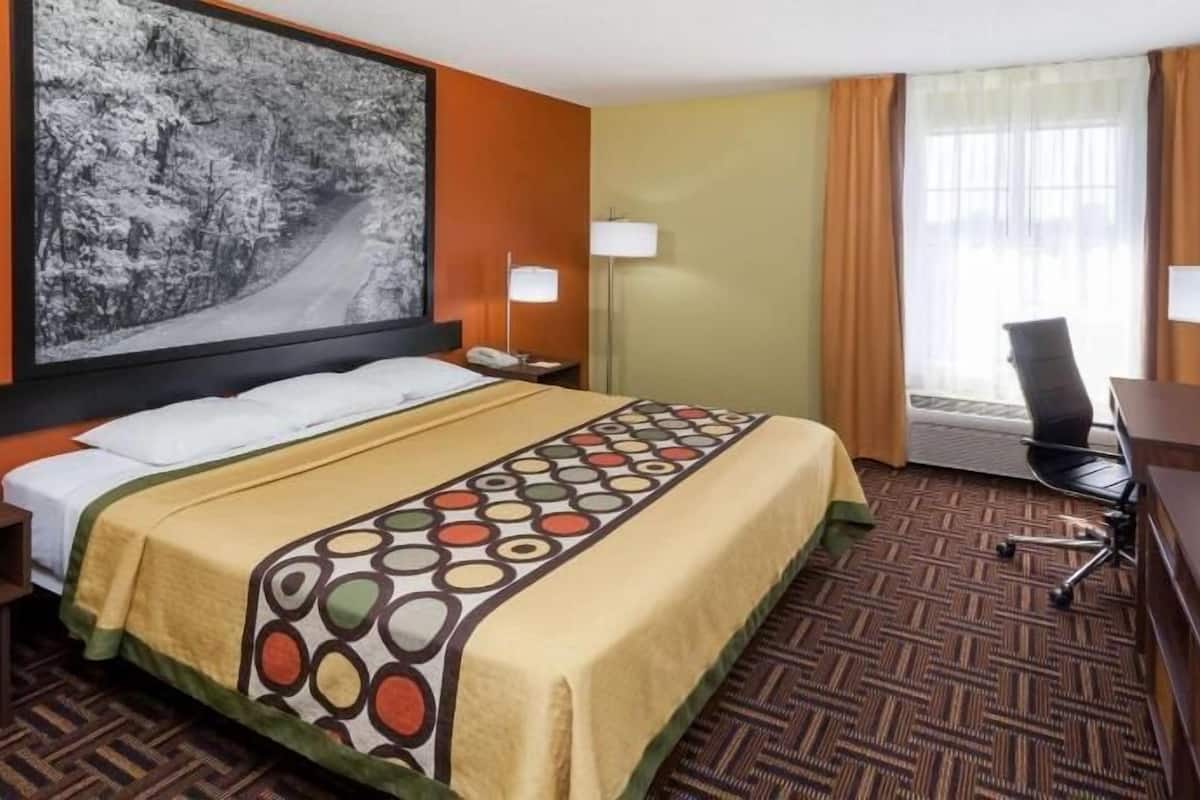 A hotel room with a bed, a desk, a chair, a TV, a large painting, and a window with curtains.