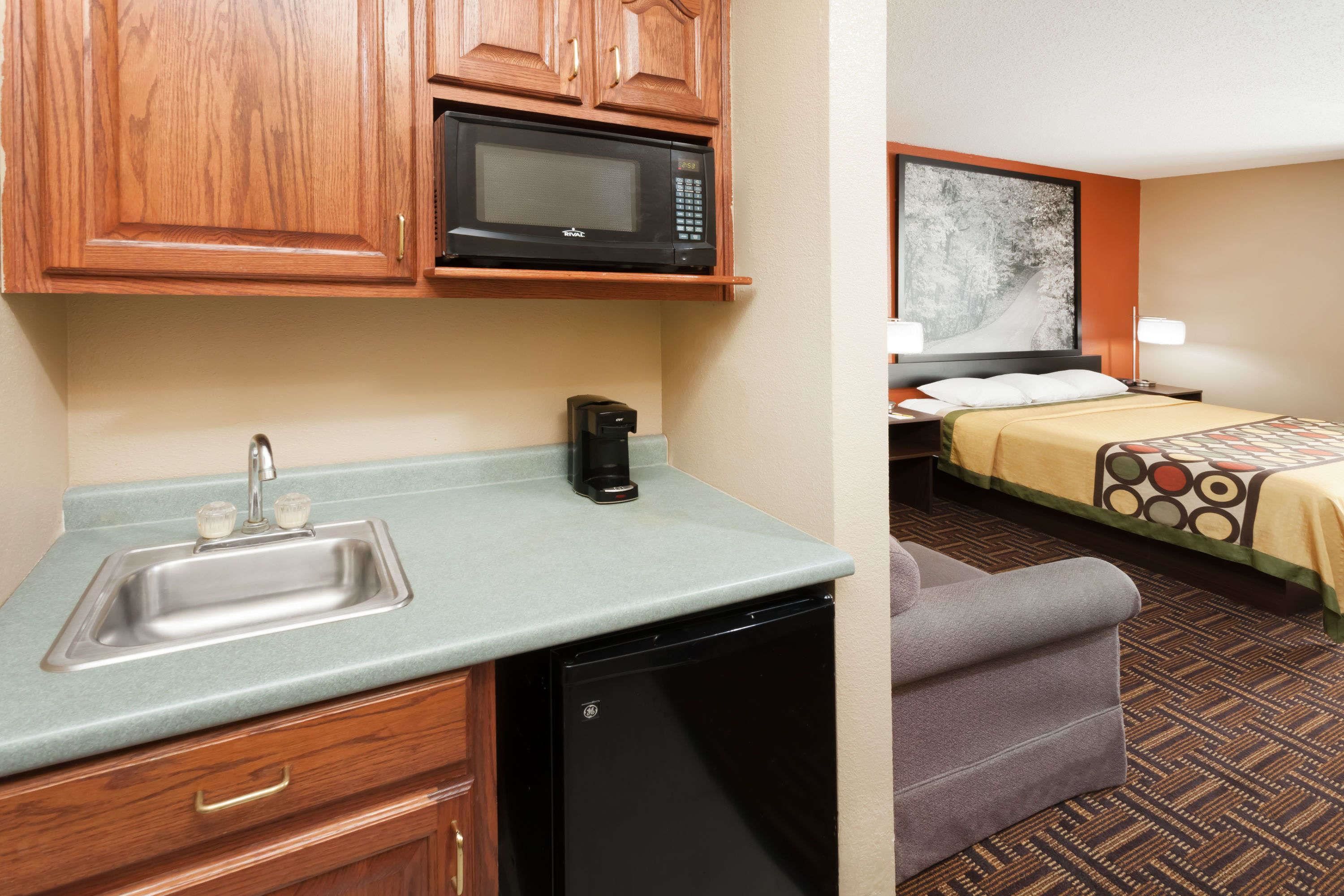 Una kitchenette equipada con microondas, lavamanos y cafetera. Se aprecia una cama con colcha estampada y un cuadro en la pared.
