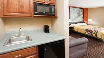 Una kitchenette equipada con microondas, lavamanos y cafetera. Se aprecia una cama con colcha estampada y un cuadro en la pared.