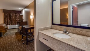 A hotel room with a bed, a desk, a chair, a large mirror, and a bathroom with a sink and toilet.
