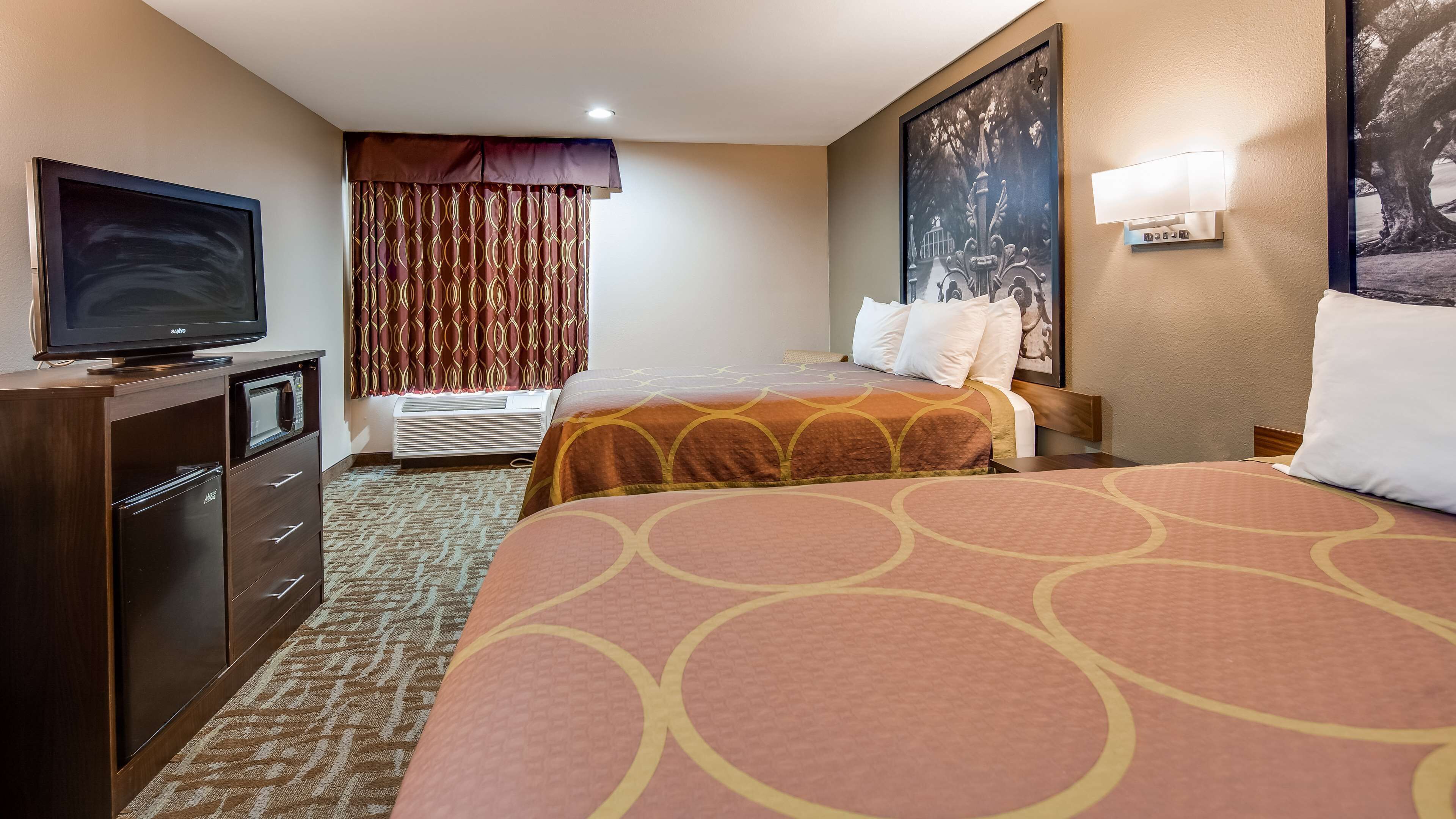 Une chambre d’hôtel équipée d’une télévision, d’un minibar, d’un lit avec une couvre-lit à motifs, d’une table de chevet avec une lampe et d’un grand tableau encadré au mur.