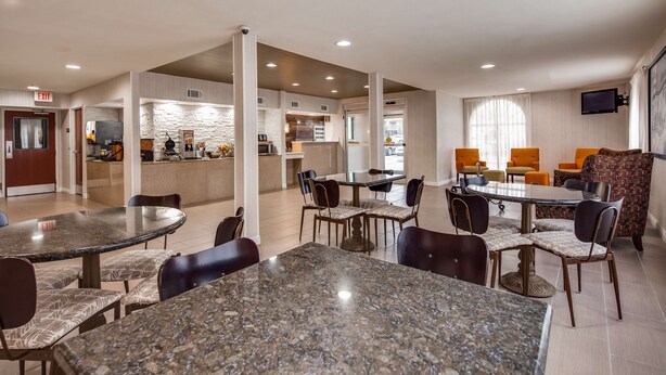 A hotel lobby with multiple dining tables and chairs, a kitchen area, and a seating area with orange chairs.