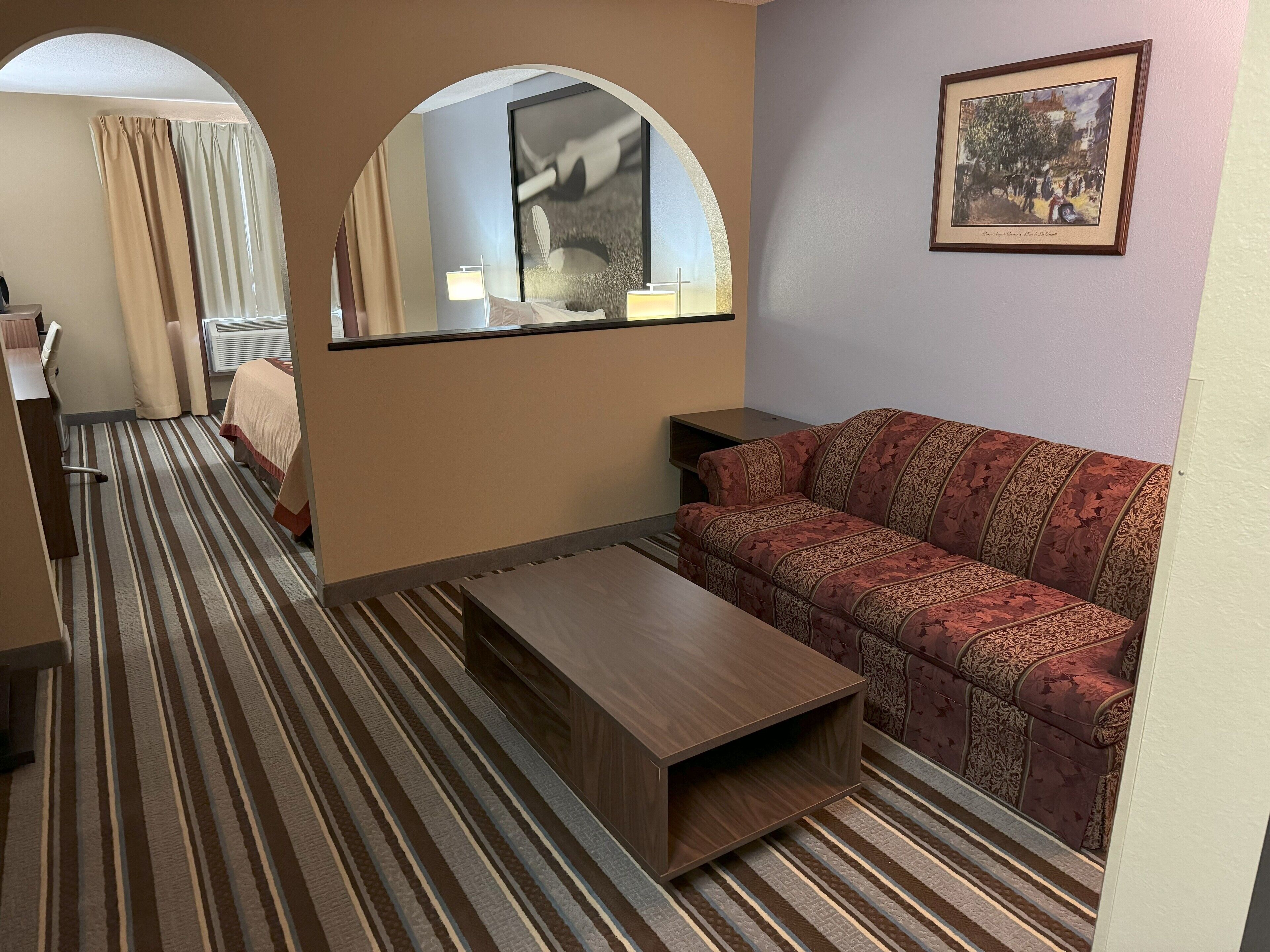 Habitació d'hotel amb una catifes rayada, un sofà, una taula de centre de fusta, un mirall, un quadre amb marc a la paret i un llit amb cortines.