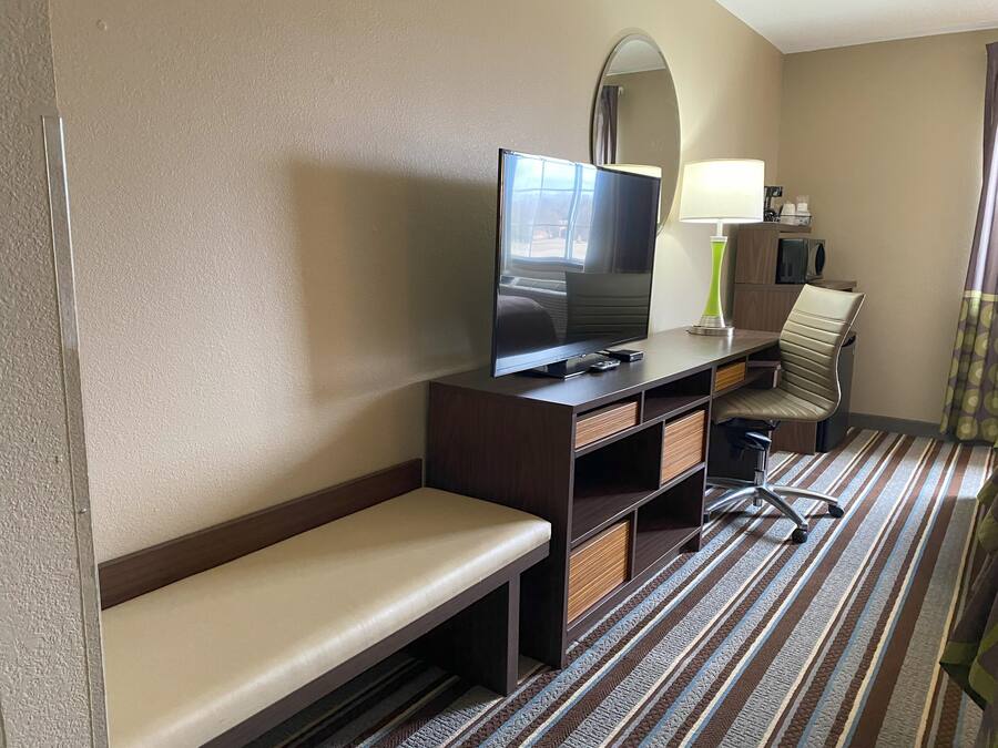 A hotel room with a flat-screen TV, a desk with a lamp, a chair, a bench, and a striped carpet.