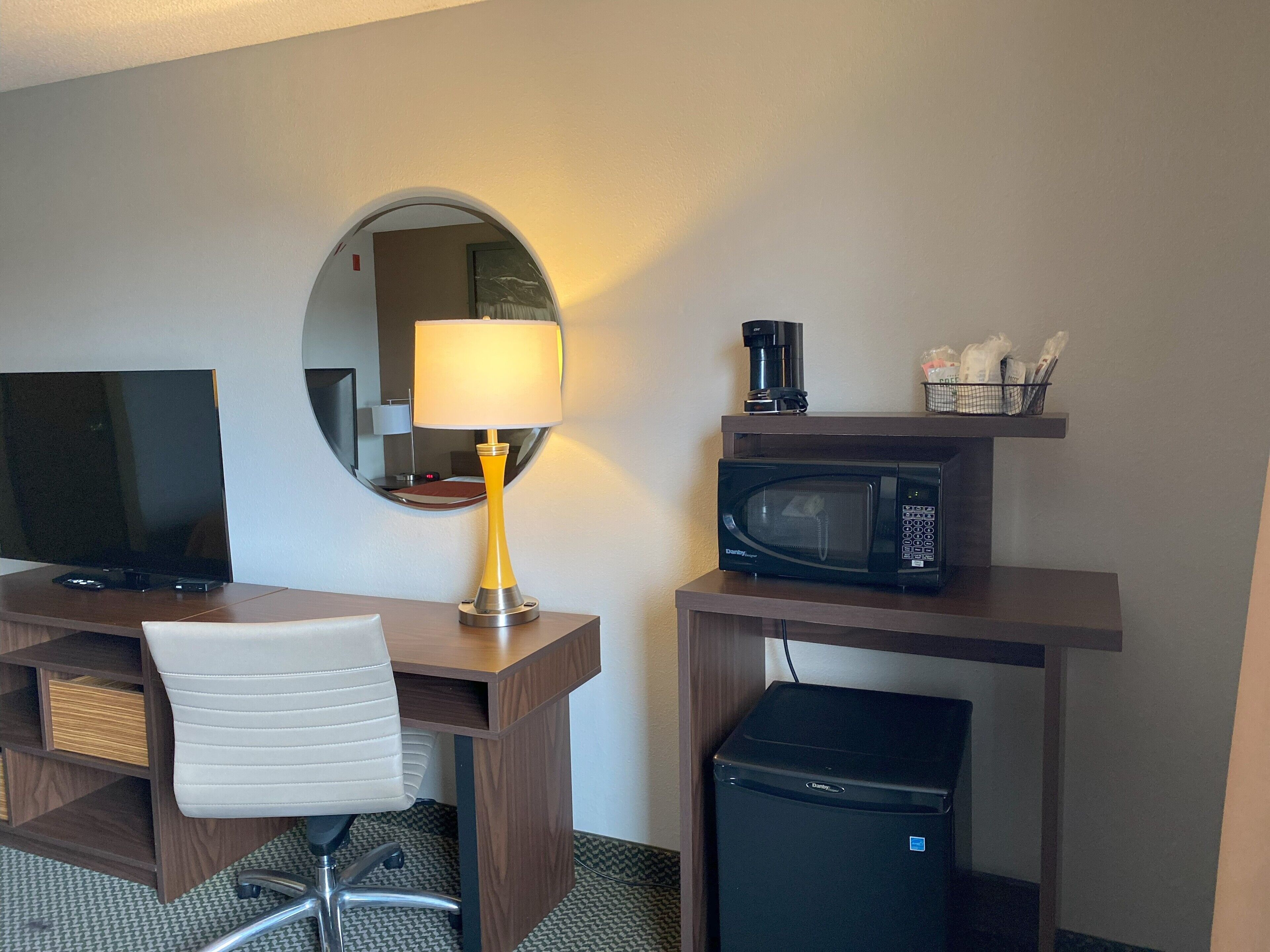 A hotel room with a desk, chair, lamp, microwave, coffee maker, and a small refrigerator.