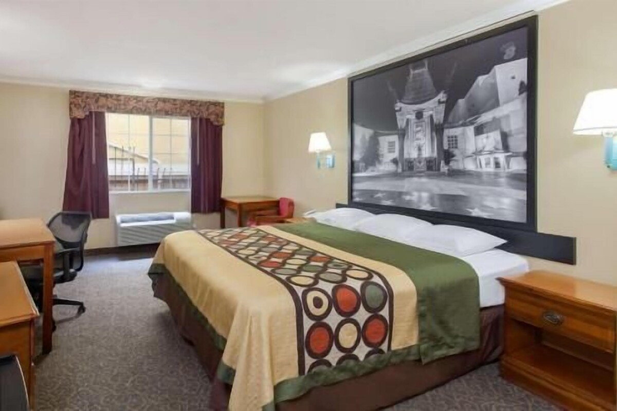 A hotel room with a bed, a desk, a chair, a window with curtains, and a wall-mounted lamp.