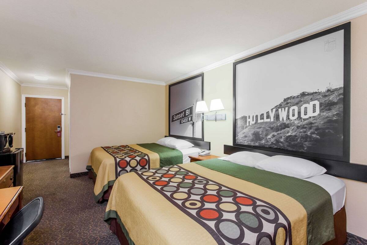 A hotel room with two beds, a desk, a chair, a lamp, and a framed picture of Hollywood.