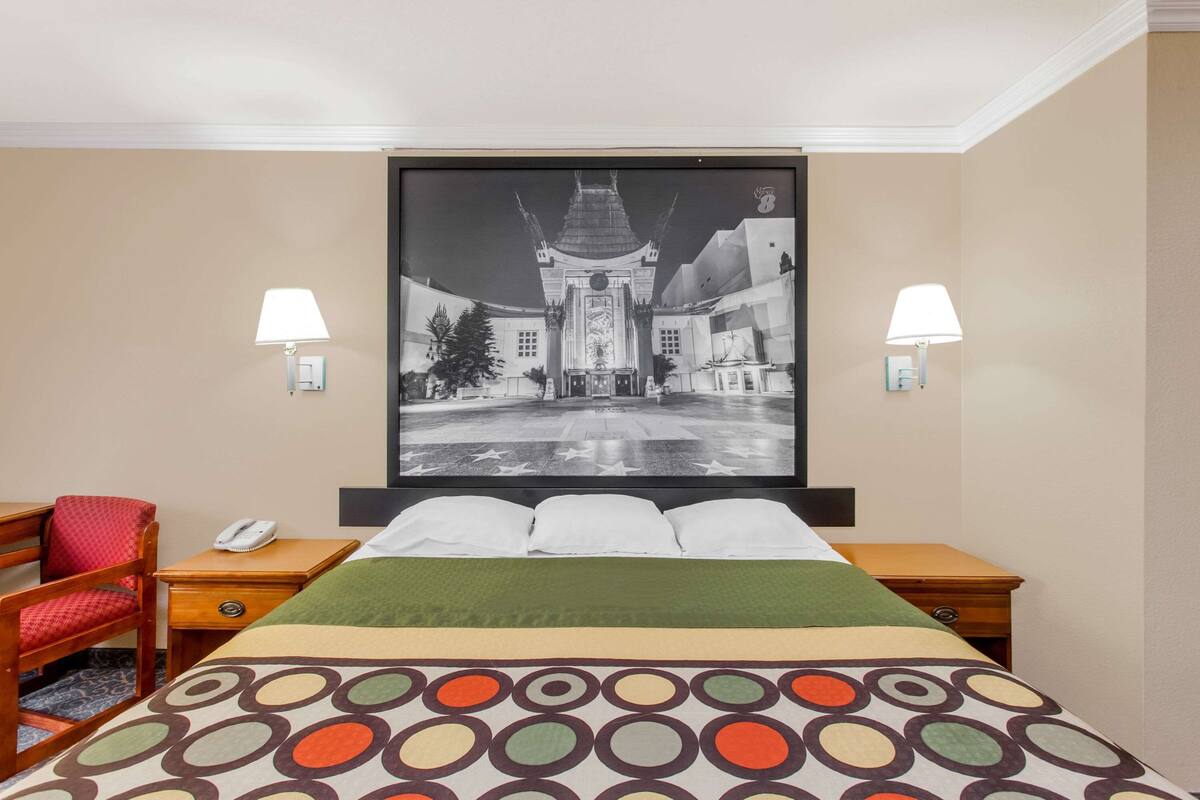 Een hotelkamer met een bed, twee nachtkastjes, een stoel, een groot gepresenteerd schilderij aan de muur en twee wandlampen.