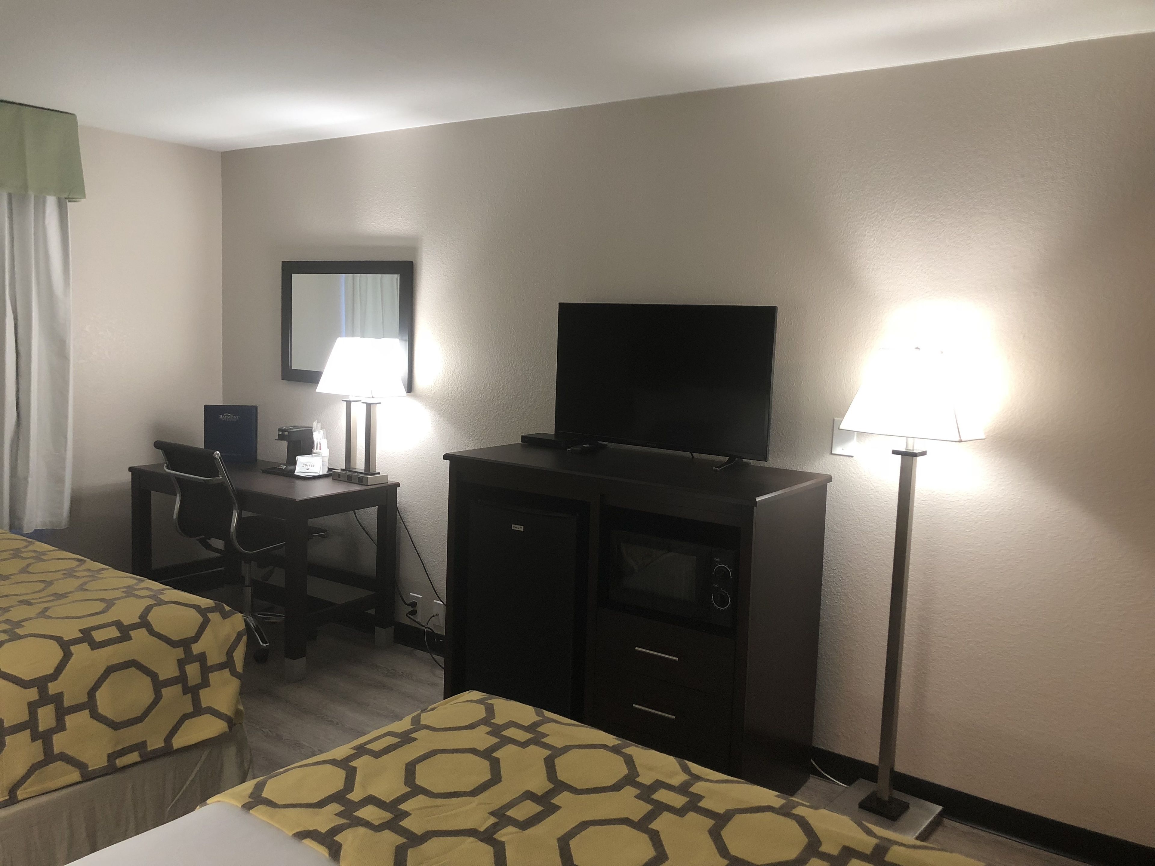 A hotel room with two beds, a desk with a computer and lamp, a TV, and a minibar.